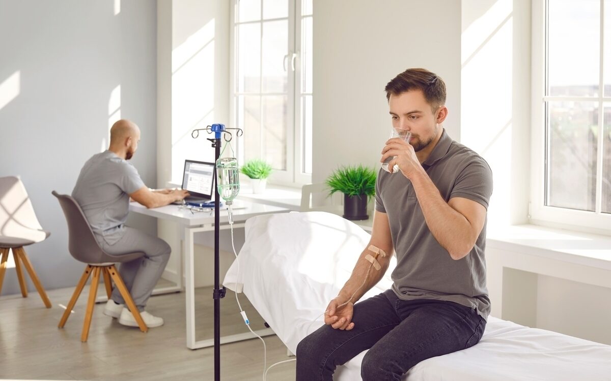 IV hydration vs drinking water comparison showing a man drinking water while receiving IV fluids in a medical clinic.