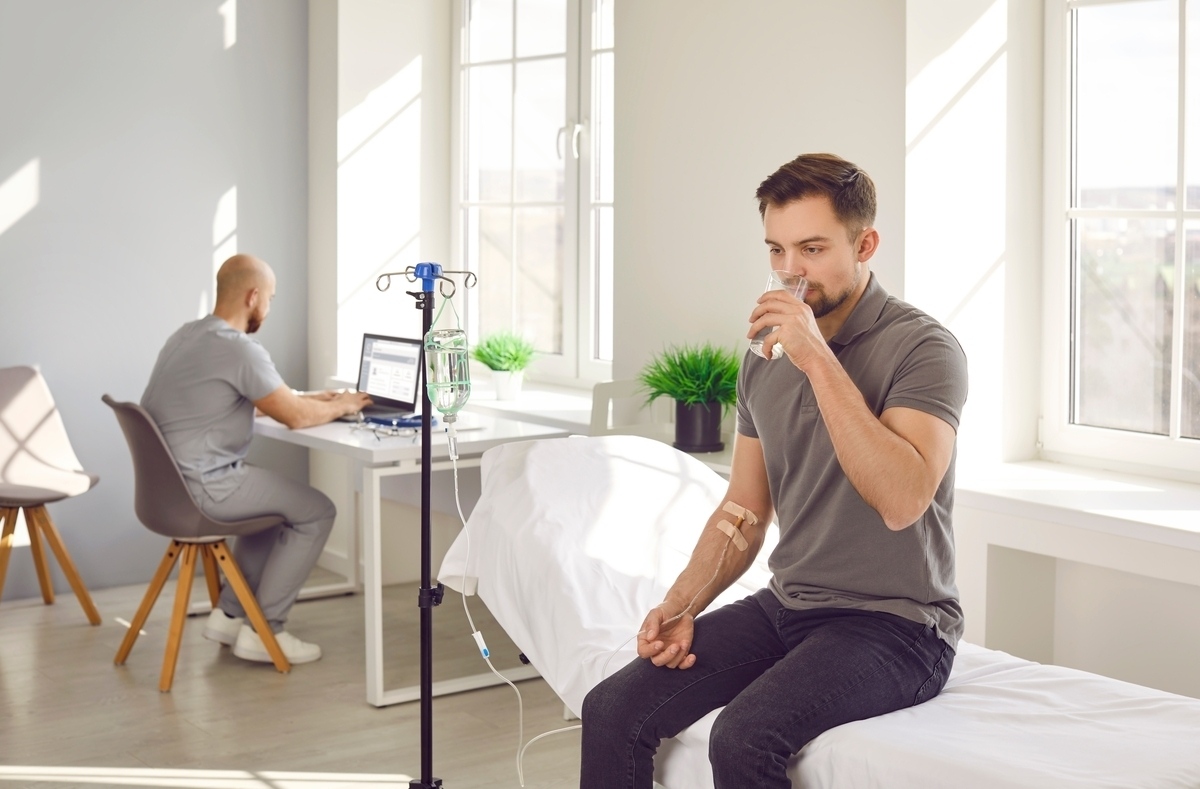 IV hydration vs drinking water comparison showing a man drinking water while receiving IV fluids in a medical clinic.
